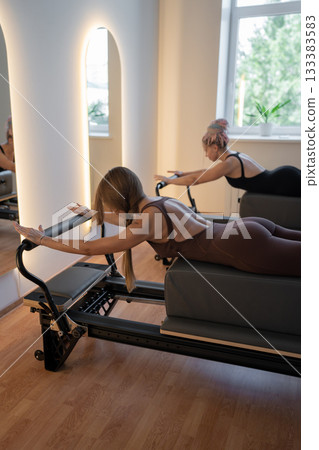 Group of caucasian women exercising with trainer on pilates reformers in a bright studio. Their workout focuses on core strength and flexibility in a modern fitness environment 133383583