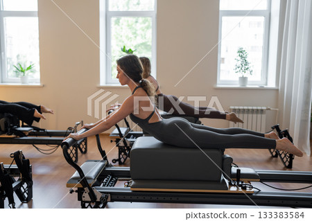 Group of caucasian women exercising with trainer on pilates reformers in a bright studio. Their workout focuses on core strength and flexibility in a modern fitness environment 133383584