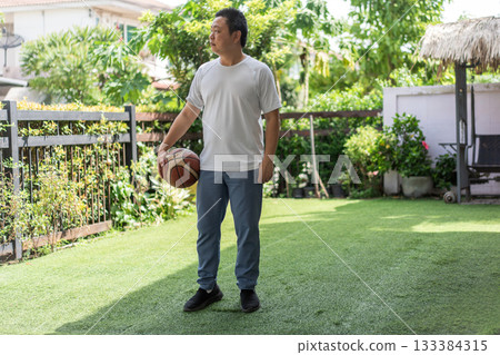 A man in a white T-shirt stands on green grass holding a basketball, relaxed and ready to move. Morning sunlight gives the scene a calm, natural energy. 133384315