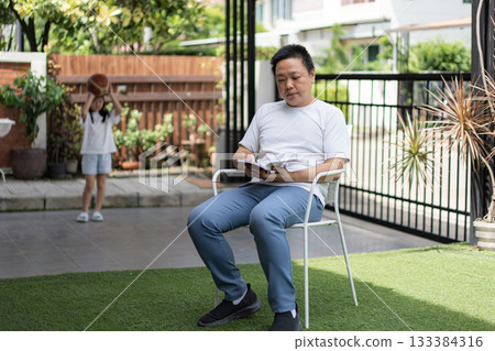 A man reads peacefully while a young girl holds a basketball in the background. A soft moment showing connection across generations at home. A man reads peacefully while a young girl holds a basketball in the background. A soft moment showing connection across generations at home. 133384316