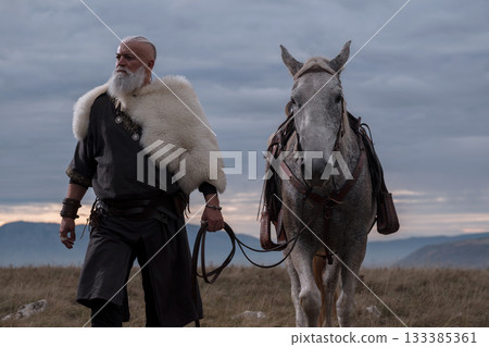 A majestic Viking chieftain on a dappled grey horse, clad in a fur cloak, surveys a desolate, mountainous landscape under a dramatic cloudy sky, exuding power and presence. A majestic Viking chieftain on a dappled grey horse, clad in a fur cloak, surveys a desolate, mountainous landscape under a dramatic cloudy sky, exuding power and presence. 133385361