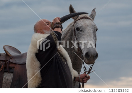 A rugged Viking man with a white beard, dressed in traditional attire, drinking from a horn against a stunning sunset background, showcasing strength, heritage, and heroic spirit A rugged Viking man with a white beard, dressed in traditional attire, drinking from a horn against a stunning sunset background, showcasing strength, heritage, and heroic spirit 133385362
