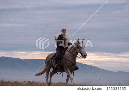 A majestic Viking chieftain on a dappled grey horse, clad in a fur cloak, surveys a desolate, mountainous landscape under a dramatic cloudy sky, exuding power and presence. 133385365
