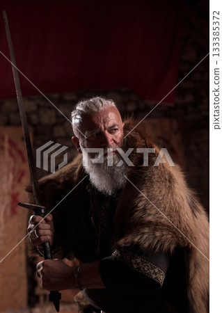 A stern Viking chieftain with a formidable white beard and braided hair, wearing a fur cloak, in battle position, ready for attack, holding a long sword, showcasing his warrior spiri A stern Viking chieftain with a formidable white beard and braided hair, wearing a fur cloak, in battle position, ready for attack, holding a long sword, showcasing his warrior spiri 133385372