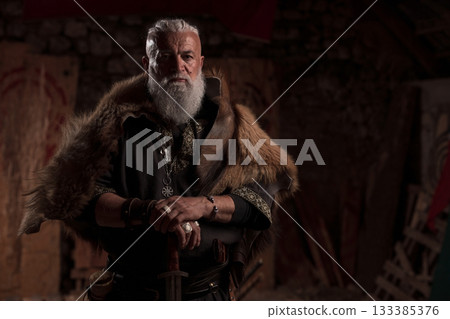 A rugged Viking man with a white beard, wearing traditional armor and a fur cloak, stands inside a stone hall, holding a sword with a contemplative expression, evoking strength and heritage. 133385376