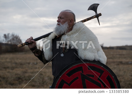 Viking warrior with axe standing on mountain cliff, wearing fur cloak and leather armor, overlooking dramatic landscape. Historical Nordic fighter 133385380