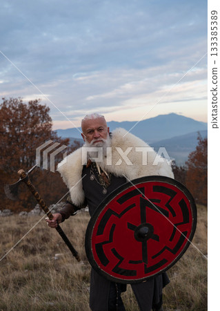 Viking warrior with axe standing on mountain cliff, wearing fur cloak and leather armor, overlooking dramatic landscape. Historical Nordic fighter 133385389