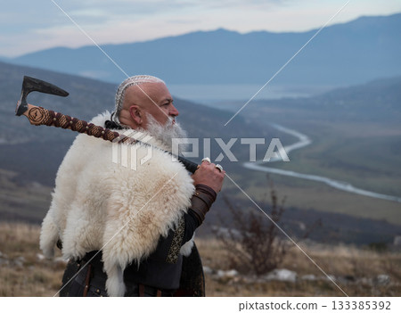 Viking warrior with axe standing on mountain cliff, wearing fur cloak and leather armor, overlooking dramatic landscape. Historical Nordic fighter 133385392
