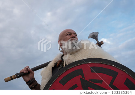 Viking warrior with axe standing on mountain cliff, wearing fur cloak and leather armor, overlooking dramatic landscape. Historical Nordic fighter 133385394