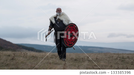 Viking warrior with axe standing on mountain cliff, wearing fur cloak and leather armor, overlooking dramatic landscape. Historical Nordic fighter 133385396