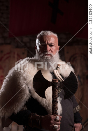 Rugged Viking warrior as a king or hero with a white beard, dressed in traditional armor, holding a battle axe, inside a medieval hall, intense and fierce, embodying strength and heritage 133385400