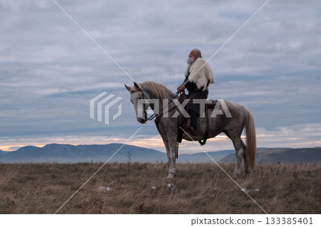 A majestic Viking chieftain on a dappled grey horse, clad in a fur cloak, surveys a desolate, mountainous landscape under a dramatic cloudy sky, exuding power and presence. A majestic Viking chieftain on a dappled grey horse, clad in a fur cloak, surveys a desolate, mountainous landscape under a dramatic cloudy sky, exuding power and presence. 133385401