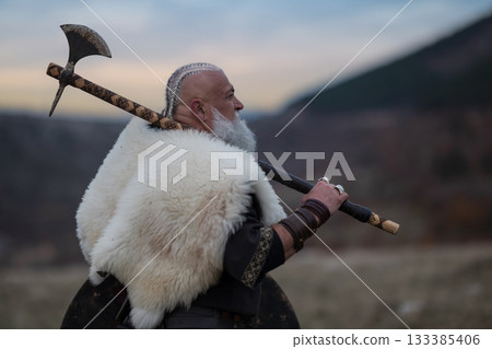 Viking warrior with axe standing on mountain cliff, wearing fur cloak and leather armor, overlooking dramatic landscape. Historical Nordic fighter 133385406
