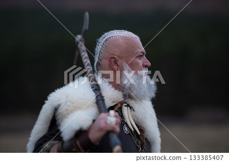 Viking warrior with axe standing on mountain cliff, wearing fur cloak and leather armor, overlooking dramatic landscape. Historical Nordic fighter 133385407