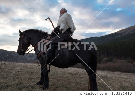 A fierce Viking warrior on horseback, wielding an axe, stands against a dramatic sunset sky with clouds and mountains, embodying strength, exploration, and ancient legend. 133385410