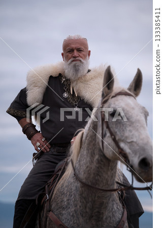 A majestic Viking chieftain on a dappled grey horse, clad in a fur cloak, surveys a desolate, mountainous landscape under a dramatic cloudy sky, exuding power and presence. A majestic Viking chieftain on a dappled grey horse, clad in a fur cloak, surveys a desolate, mountainous landscape under a dramatic cloudy sky, exuding power and presence. 133385411