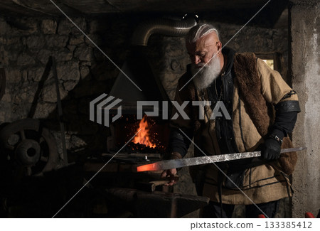 A Viking blacksmith with a white beard and braided hair works intently at a forge, hammering glowing metal on an anvil, with flames flickering in the background of a stone workshop. 133385412