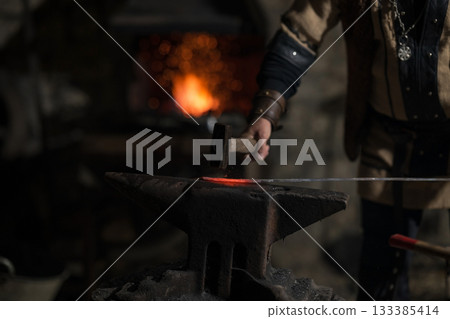 A Viking blacksmith with a white beard and braided hair works intently at a forge, hammering glowing metal on an anvil, with flames flickering in the background of a stone workshop. 133385414