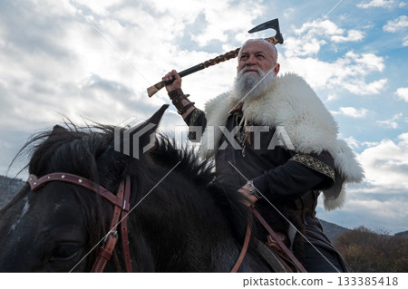 A fierce Viking warrior on horseback, wielding an axe, stands against a dramatic sunset sky with clouds and mountains, embodying strength, exploration, and ancient legend. 133385418