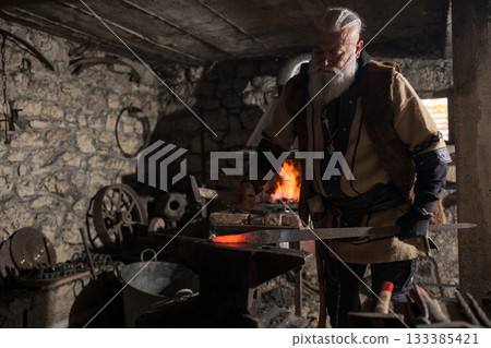 A Viking blacksmith with a white beard and braided hair works intently at a forge, hammering glowing metal on an anvil, with flames flickering in the background of a stone workshop. 133385421