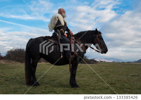 A Viking warrior on a large black horse surveys a dramatic landscape during sunset, wearing a fur cloak. The scene evokes ancient exploration, adventure, and the spirit of the wild. 133385426
