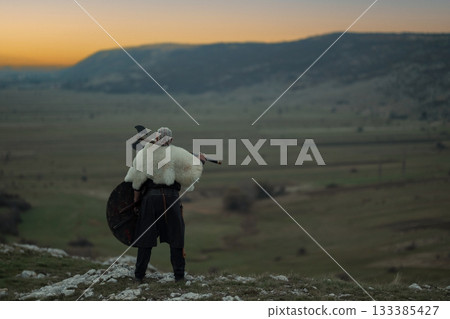 Viking warrior with axe standing on mountain cliff, wearing fur cloak and leather armor, overlooking dramatic landscape. Historical Nordic fighter 133385427