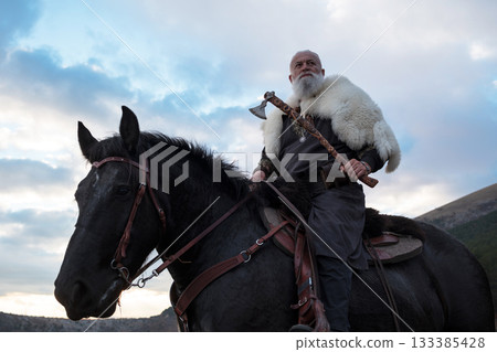 A fierce Viking warrior on horseback, wielding an axe, stands against a dramatic sunset sky with clouds and mountains, embodying strength, exploration, and ancient legend. 133385428