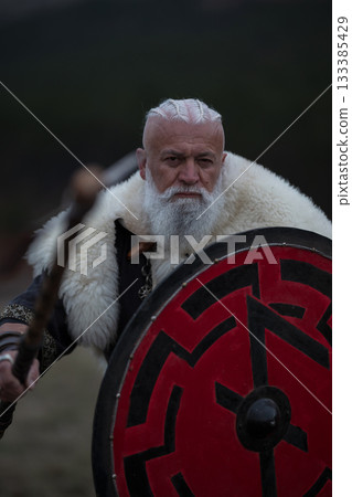 Viking warrior with axe standing on mountain cliff, wearing fur cloak and leather armor, overlooking dramatic landscape. Historical Nordic fighter 133385429