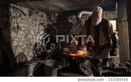 A Viking blacksmith with a white beard and braided hair works intently at a forge, hammering glowing metal on an anvil, with flames flickering in the background of a stone workshop. 133385430