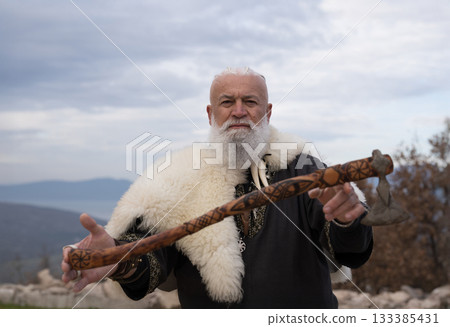 Viking warrior with axe standing on mountain cliff, wearing fur cloak and leather armor, overlooking dramatic landscape. Historical Nordic fighter 133385431