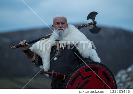 Viking warrior with axe standing on mountain cliff, wearing fur cloak and leather armor, overlooking dramatic landscape. Historical Nordic fighter Viking warrior with axe standing on mountain cliff, wearing fur cloak and leather armor, overlooking dramatic landscape. Historical Nordic fighter 133385436