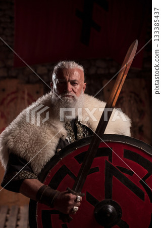 A stern Viking chieftain with a formidable white beard and braided hair, wearing a fur cloak, gazes directly at the viewer while holding a long sword upright, showcasing his warrior spirit. 133385437