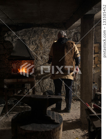 A Viking blacksmith with a white beard and braided hair works intently at a forge, hammering glowing metal on an anvil, with flames flickering in the background of a stone workshop. 133385452