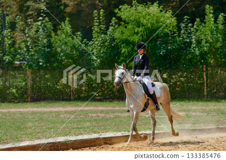 Equestrian sport, horse and rider in dressage competition, woman riding horse in outdoor arena, training and performance, equine event, sunny day, natural light, action shot. Equestrian sport, horse and rider in dressage competition, woman riding horse in outdoor arena, training and performance, equine event, sunny day, natural light, action shot. 133385476