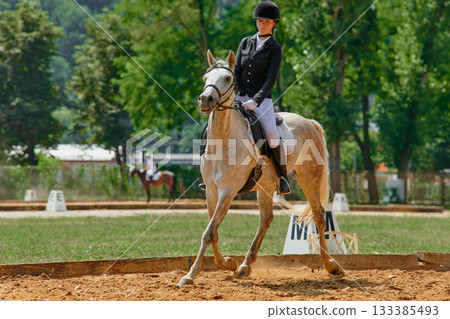 Equestrian sport, horse and rider in dressage competition, woman riding horse in outdoor arena, training and performance, equine event, sunny day, natural light, action shot. Equestrian sport, horse and rider in dressage competition, woman riding horse in outdoor arena, training and performance, equine event, sunny day, natural light, action shot. 133385493