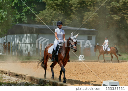 Equestrian sport, young woman riding a horse in dressage competition, outdoor training, horse riding school, equestrian center, horse and rider in action, sunny day, outdoor scene. Equestrian sport, young woman riding a horse in dressage competition, outdoor training, horse riding school, equestrian center, horse and rider in action, sunny day, outdoor scene. 133385531
