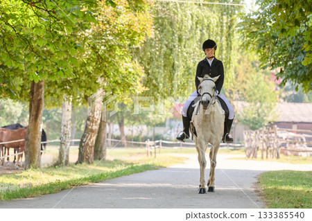 Woman riding a white horse on a country road, equestrian sport, horse riding, outdoor activity, summer landscape, countryside scene, equine lifestyle, animal portrait. Woman riding a white horse on a country road, equestrian sport, horse riding, outdoor activity, summer landscape, countryside scene, equine lifestyle, animal portrait. 133385540
