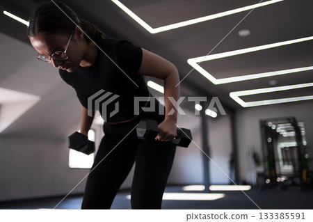 Woman exercises with dumbbells in a modern gym during daylight hours focusing on strength training Woman exercises with dumbbells in a modern gym during daylight hours focusing on strength training 133385591