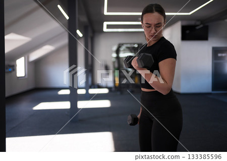 Woman lifts dumbbell during workout in modern gym with natural light in the afternoon Woman lifts dumbbell during workout in modern gym with natural light in the afternoon 133385596