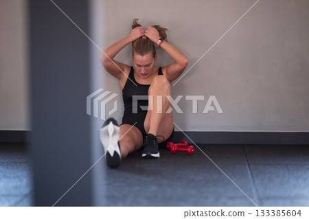 Woman exercising alone indoors sits against the wall after a workout session with dumbbells at a gym 133385604