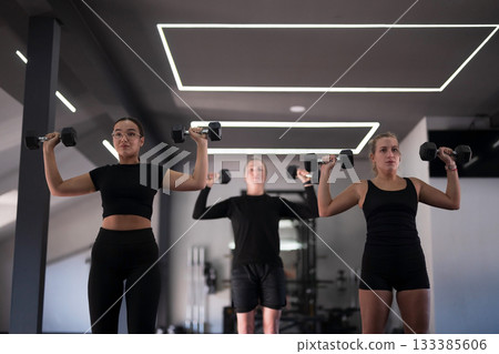 Women workout together lifting dumbbells in a modern gym during a fitness class session Women workout together lifting dumbbells in a modern gym during a fitness class session 133385606