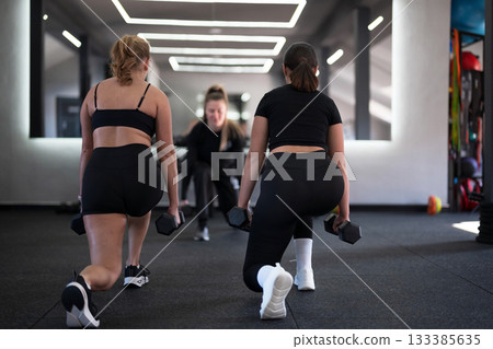 Two women performing lunges with dumbbells during a workout session in a modern gym environment Two women performing lunges with dumbbells during a workout session in a modern gym environment 133385635