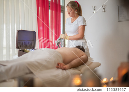 Professional female therapist wearing gloves and preparing equipment while a man lies on a massage table covered with a towel. Spa and wellness treatment concept highlighting relaxation, health care 133385637