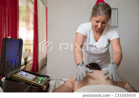 Close up view of a therapist applying electrotherapy with special conductive gloves on a man back during a physiotherapy session. Concept of modern rehabilitation, pain relief, muscle stimulation, and Close up view of a therapist applying electrotherapy with special conductive gloves on a man back during a physiotherapy session. Concept of modern rehabilitation, pain relief, muscle stimulation, and 133385650