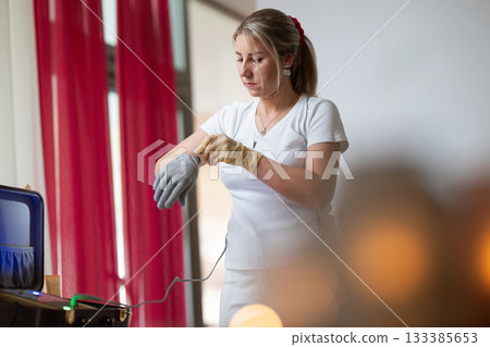 Professional female therapist wearing gloves and preparing equipment while a man lies on a massage table covered with a towel. Spa and wellness treatment concept highlighting relaxation, health care 133385653