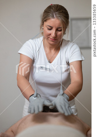 Close up view of a therapist applying electrotherapy with special conductive gloves on a man back during a physiotherapy session. Concept of modern rehabilitation, pain relief, muscle stimulation, and 133385659