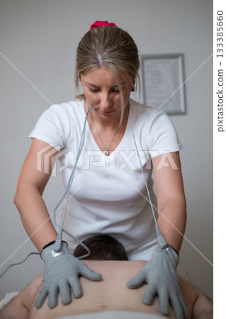 Close up view of a therapist applying electrotherapy with special conductive gloves on a man back during a physiotherapy session. Concept of modern rehabilitation, pain relief, muscle stimulation, and Close up view of a therapist applying electrotherapy with special conductive gloves on a man back during a physiotherapy session. Concept of modern rehabilitation, pain relief, muscle stimulation, and 133385660