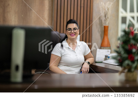 Female receptionist wearing glasses and white uniform smiling at the front desk of a spa and wellness center, representing hospitality, customer care, and professional service. 133385692