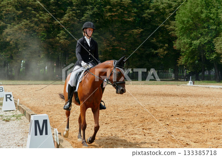 Equestrian dressage competition, young woman riding a horse in arena, outdoor training, horse show event, skill and precision, equestrian sport, sunny day. Equestrian dressage competition, young woman riding a horse in arena, outdoor training, horse show event, skill and precision, equestrian sport, sunny day. 133385718