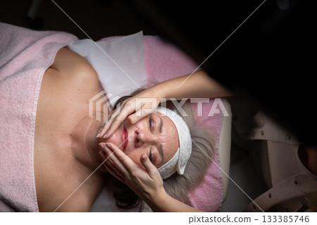 Close up of a mature woman receiving a relaxing facial massage from a professional female therapist in a modern spa. The client lies on a treatment bed with a towel and headband, enjoying skincare Close up of a mature woman receiving a relaxing facial massage from a professional female therapist in a modern spa. The client lies on a treatment bed with a towel and headband, enjoying skincare 133385746